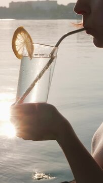 Anonymous woman drinks cold water with slice of lemon through reusable metal tube in hot weather. Brunette holding tall glass stands on seashore, on which surface sun's rays are reflected