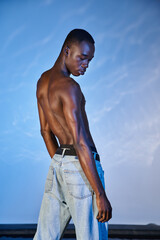 alluring young african american man in stylish jeans posing topless on watery blue background