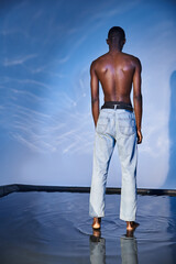 back view of fashionable shirtless african american man in trendy jeans on blue watery backdrop