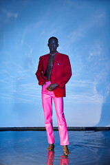 alluring stylish african american man in pink pants and red blazer posing on blue watery backdrop