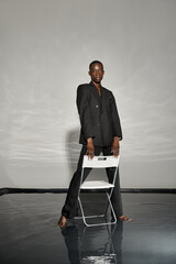 charming african american man in sexy black suit posing next to chair on gray watery backdrop