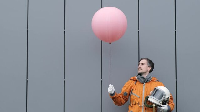 Hero astronaut wearing space suit holding pink helium balloon against gray wall. 4k surreal concept
