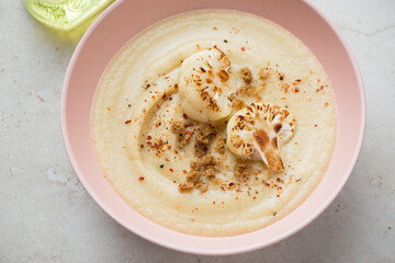 Cream-soup made of roasted cauliflower and served in a roseate bowl, horizontal shot on a light-beige stone surface, middle closeup