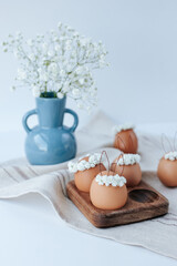On a linen napkin there is a wooden egg cup with brown Easter eggs with flower wreaths and bunny ears. In the background there is a blue vase with gypsophila flowers. White background