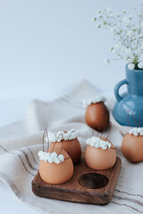 On a linen napkin there is a wooden egg cup with brown Easter eggs with flower wreaths and bunny ears. In the background there is a blue vase with gypsophila flowers. White background
