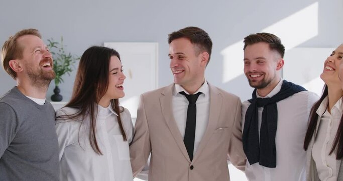 Group of laughing business people men and women looking at camera hugging and smiling indoors. Company employees talking standing in office. Team work concept. 4k video. Slow motion video.