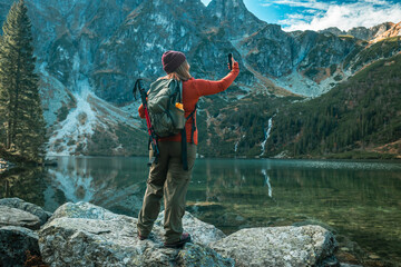 Rear shot of traveler woman influencer in sportswear with backpack standing on the rocky shore and making selfie on smart phone. Taking photo of green hills and mountains on Morskie Oko lake. Hiking - obrazy, fototapety, plakaty