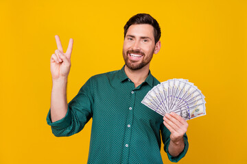 Photo of cheerful pleasant guy with beard dressed dotted shirt holding money showing v-sign symbol...