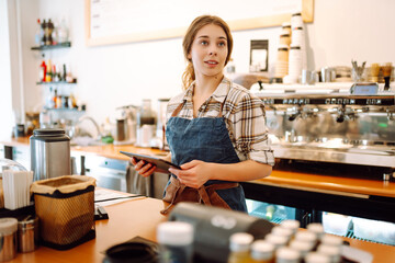 Smiling female barista takes an order from a tablet while standing at the bar counter in a coffee...