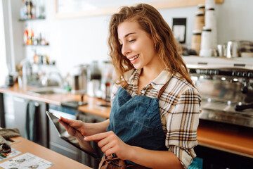 Smiling female barista takes an order from a tablet while standing at the bar counter in a coffee...