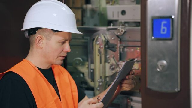 A utility worker repairs an elevator in a residential complex. A repairman in uniform is intently identifying the cause of a breakdown in a passenger elevator and fixing the problem.