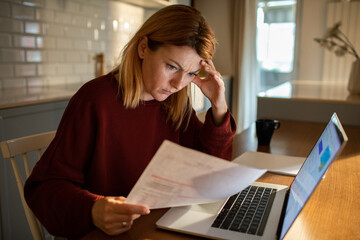 Worried woman reviewing documents with laptop at kitchen table