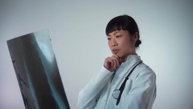 Serious Asian female doctor examining a lung radiography isolated on grey background. Doctor examining chest x-ray film of patient at hospital. Doctor diagnosing patient’s health on asthma, lung