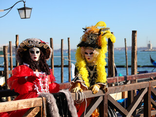 Two masked women with hats and wigs on the sea pier of Venice in Italy