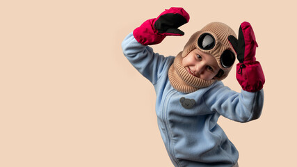 Child in warm goggle hat with mittens on hands on beige background. Copy space.
