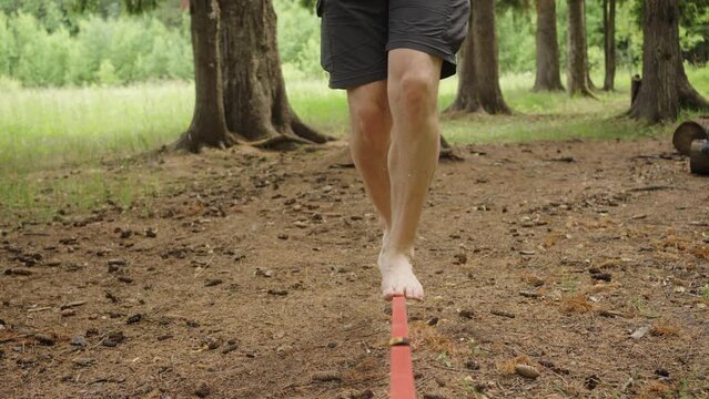 Barefoot balance on a slackline in a serene forest setting, highlighting a moment of concentration and physical coordination. A man walks along a stretched sling in a park between trees, legs in close