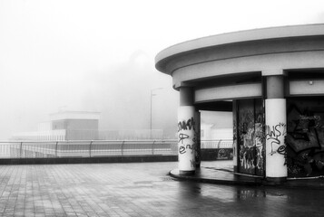 Along the mist-shrouded promenade, the outlines of buildings and lampposts blur into obscurity,...