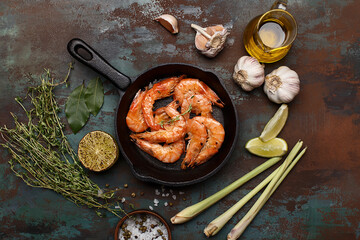 Grilled shrimps in cast iron pan with lime, lemongrass, thyme, garlic, oil and salt over dark green background. Top view. Flat composition of asian food ingredients