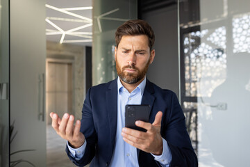 A worried young man businessman looks at the screen of the mobile phone he is holding in his hand and spreads his hands in surprise while in the office