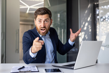 Angry young man businessman sitting in the office at the table and pointing his finger at the...