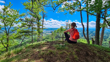Naklejka na ściany i meble Hiker woman at summit cross of Heiggerkogel in Grazer Bergland, Prealps East of the Mur, Styria, Austria. Scenic view of soft hill landscape. Idyllic hiking trail through lush green forest. Wanderlust