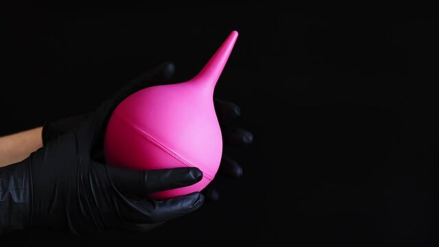 A hand in a black latex glove holds a large pink enema on a dark background. Medical device. Pear-shaped enema, body cleansing concept