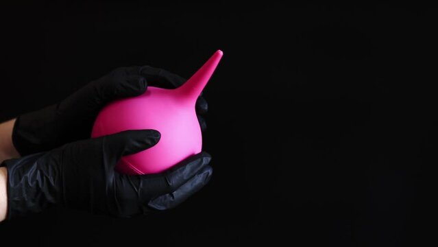 A hand in a black latex glove holds a large pink enema on a dark background. Medical device. Pear-shaped enema, body cleansing concept