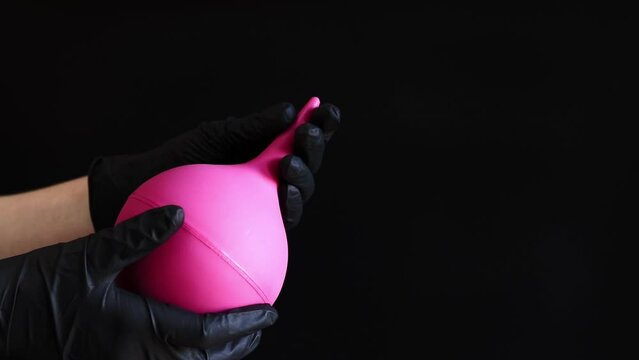 A hand in a black latex glove holds a large pink enema on a dark background. Medical device. Pear-shaped enema, body cleansing concept