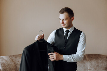 An elegant groom in a white shirt and a stylish tie is holding a jacket on a hanger. Details of the...