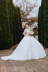 Stylish, young groom and beautiful bride in a long white dress and a long veil with a bouquet in their hands, hugging in the park in the autumn nature. Wedding portrait of newlyweds.