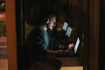 Focused man with headphones working on laptop at night in home office.