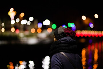 Kobieta tyłem ubrana w czapkę, szalik i kurtkę patrząca w dal na światła miasta nocą odbijające się w wodzie