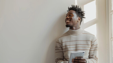 a person is casually leaning against a white wall, holding a tablet with a thoughtful smile, wearing a striped sweater.
