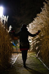 Postać kobiety tyłem idącej alejką w parku miejskim pomiędzy wysokimi trawami pampasowymi nocą