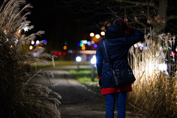 Postać kobiety tyłem idącej alejką w parku miejskim pomiędzy wysokimi trawami pampasowymi nocą - obrazy, fototapety, plakaty