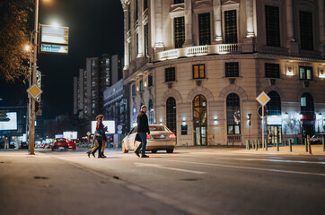 Naklejka na ściany i meble People crossing the street in a bustling city nighttime setting.