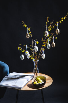interior, holidays and home decor concept - close up of easter eggs in vase with forsythia branches and magazine on table in dark room