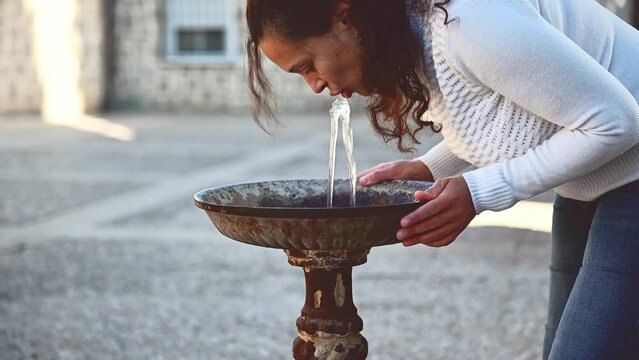 Dark-haired pretty woman drinks water flowing from a special street spring in the city. Relief from thirst in hot summer. Quesada, Jaen province, Andalusia, Spain