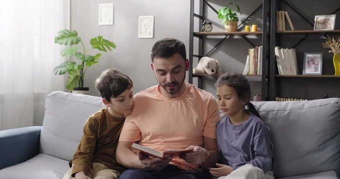 Happy family loving father reading book to his children sitting on the couch in the living room. Father, daughter and son bonding, reading interesting story, enjoy leisure time. Family tender moments