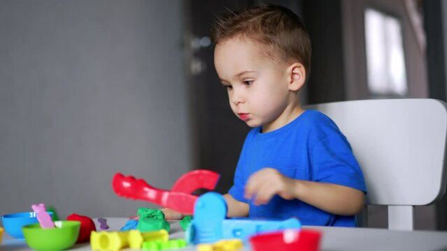 Concentrated Caucasian toddler sits at his desk at home. Cute baby boy sculpturing with plasticine.