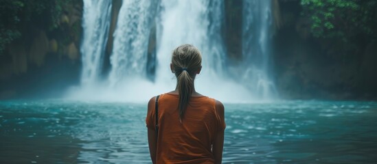 Obraz premium A woman stands facing a tall tropical waterfall, observing its cascading water with lush greenery surrounding her.