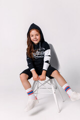 Little stylish smiling girl wearing a black hoodie sitting on white background in studio.