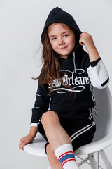 Little stylish smiling kid girl wearing a black hoodie sitting on white background in studio.