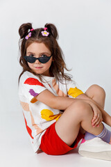 Cute little kid girl in white t-shirt and red shorts looking at camera and sitting on white studio background.