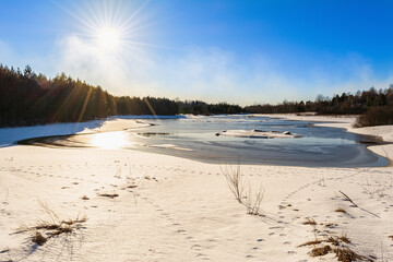 A forest lake with blue water and melting ice. Bare trunks of trees and pines on the horizon of the shoreline. View from the shore covered with snow. Spring sunny landscape