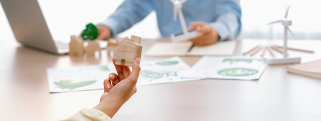 Cropped image of businesswoman presents eco-friendly house by using green design to manager while holding the wooden house block at table with windmill model placed with document. Delineation.