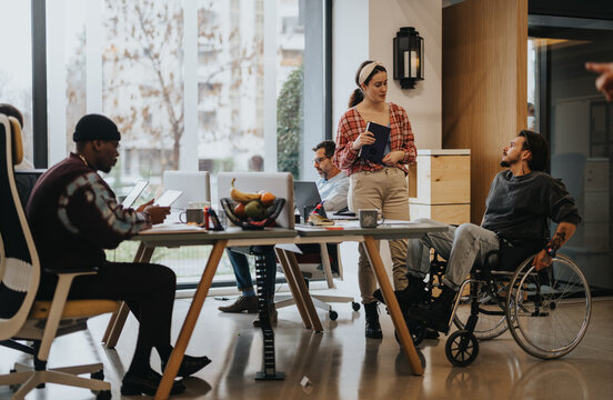 Diverse office team working and communicating in an accessible workspace. Some team members are using laptops while one is in a wheelchair, highlighting inclusivity.