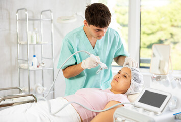 Young male cosmetologist performs hardware facial moisturizing procedure to young female patient