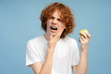 Sad, red haired curly teenager with dental braces, hand on cheek, expressing agony from apple bite