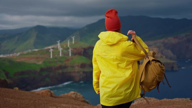 Attractive traveling woman in a yellow jacket and backpack looking at windmills in field 